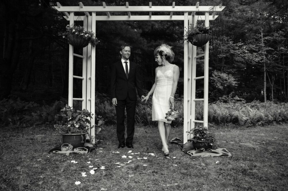 A black-and-white photograph of a couple walking under a wooden archway decorated with hanging plants and foliage. The man is wearing a suit and tie, and the woman is wearing a sleeveless dress and a headpiece. Flower petals are scattered on the grass beneath them.
