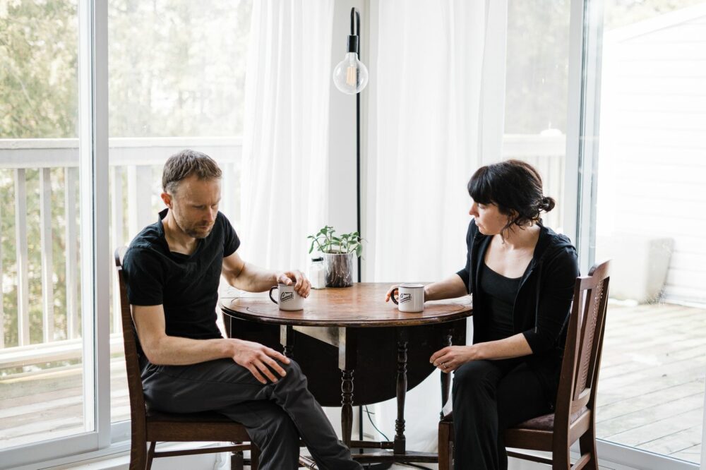 A man and woman sit across from each other at a small round table in a bright room with large glass doors. A potted plant and two coffee mugs are on the table. They are both casually dressed in dark clothing and appear to be in conversation.
