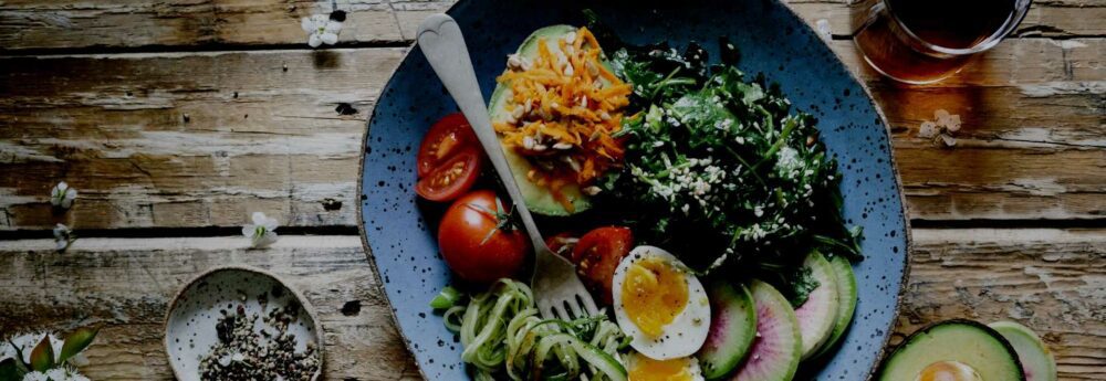 A plate of food on a wooden table. The plate contains a salad with greens, sliced cucumbers, halved cherry tomatoes, a boiled egg, shredded carrots, and a fork. There is a small dish with pepper and a glass of dark colored beverage beside the plate.