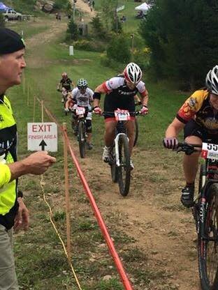 A group of cyclists is riding on a dirt trail during a race. An official wearing a yellow vest and holding a megaphone stands next to a sign reading "EXIT Finish Area." The cyclists are wearing helmets and numbered jerseys. Trees and a grassy area are in the background.
