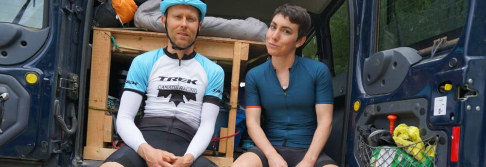 Two individuals in cycling attire sit in the open back of a van. The person on the left wears a blue helmet and a white and blue cycling jersey, while the person on the right wears a dark blue jersey. A third person is lying down inside the van.