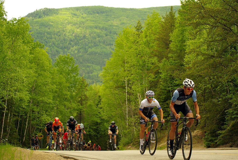 A group of cyclists rides uphill on a forested road with green trees and a mountainous backdrop. The cyclists are wearing race gear and helmets. The lead cyclist is in a black kit and another cyclist in a white kit follows closely behind.