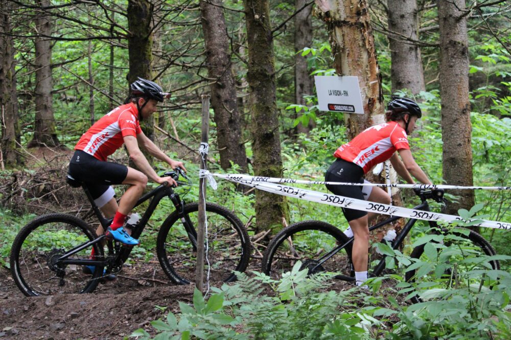 Two cyclists in red jerseys and helmets ride mountain bikes on a forest trail. The trail is bordered by white tape marked "UCI" and "JSO." One cyclist is ahead, while the other is just behind. Lush greenery and trees surround the path.