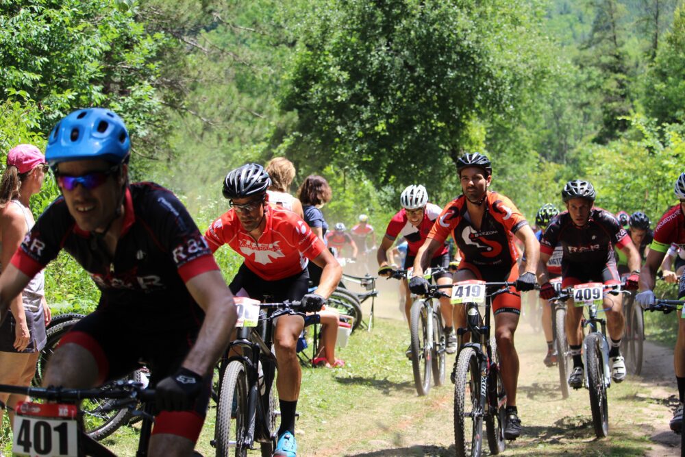A group of cyclists, all wearing helmets and numbered jerseys, ride mountain bikes on a dirt trail surrounded by trees and greenery. Spectators stand nearby, watching the race. Dust is visible in the air as the cyclists move forward.