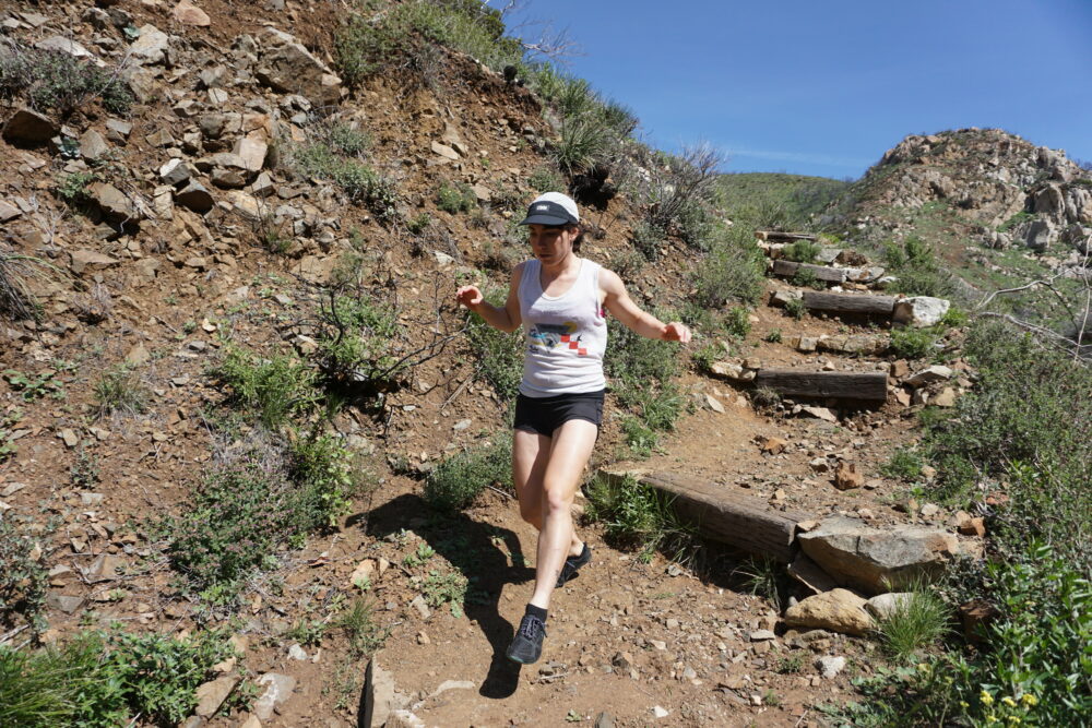 A person wearing a white tank top, black shorts, and a cap, is running downhill on a rocky trail in a mountainous, arid landscape. There is sparse vegetation around, and the sky is clear and blue.