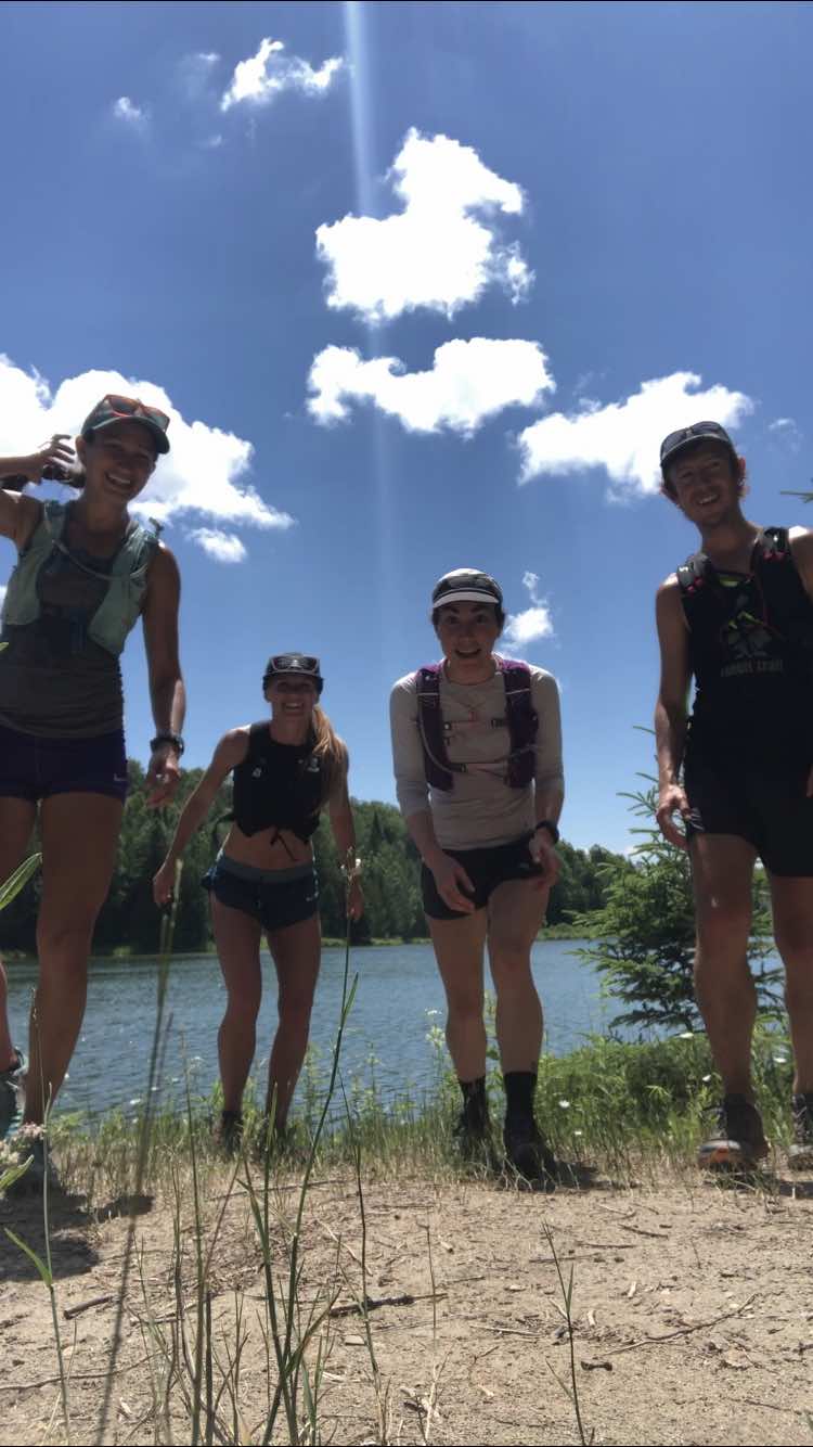 Four friends posing happily at the edge of a lake under a clear sky. They are dressed in sportswear, ready for an outdoor adventure, with lush greenery and water in the background.