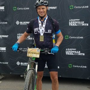 A cyclist wearing a helmet and cycling gear stands with his bike in front of a backdrop that reads "FINISHER." He has a medal around his neck and a bib number on his bike's handlebars. The backdrop features logos for Lifetime, CenturyLink, and BLUERPRINT Leadingville Trail 100 MTB.