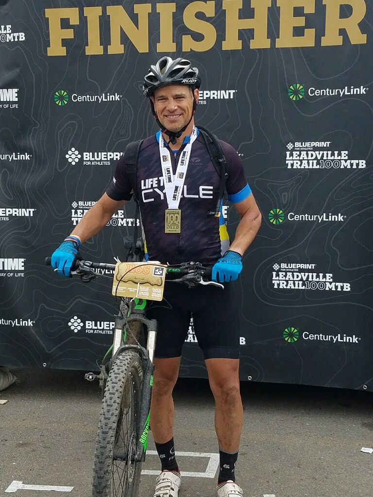 A cyclist wearing a helmet and cycling gear stands with his bike in front of a backdrop that reads "FINISHER." He has a medal around his neck and a bib number on his bike's handlebars. The backdrop features logos for Lifetime, CenturyLink, and BLUERPRINT Leadingville Trail 100 MTB.