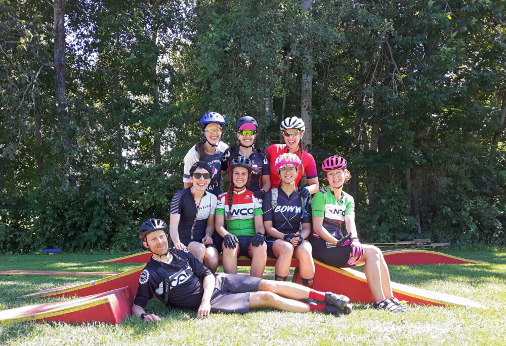 A group of eight cyclists wearing helmets and cycling attire pose for a photo in an outdoor park. Three individuals stand at the back, while five are seated or reclined on a red structure on the grass. Trees and greenery are visible in the background.