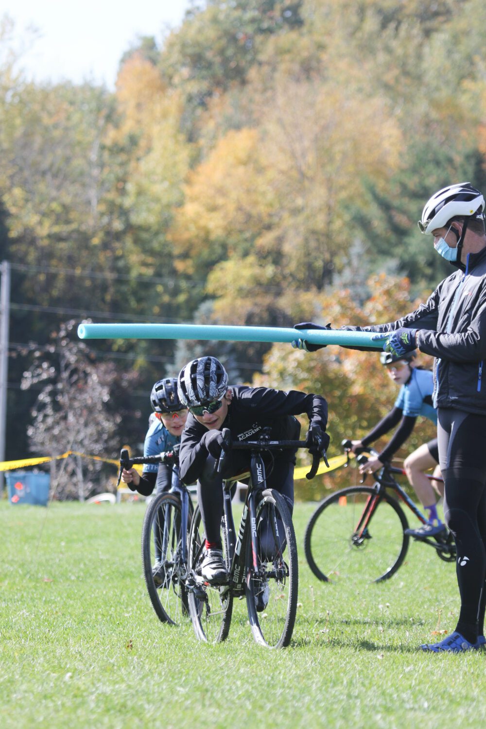 Cyclists participate in a race on grass while a person wearing a mask and holding a foam pool noodle stands nearby. The cyclists are bent over their handlebars, and trees with autumn foliage are visible in the background.
