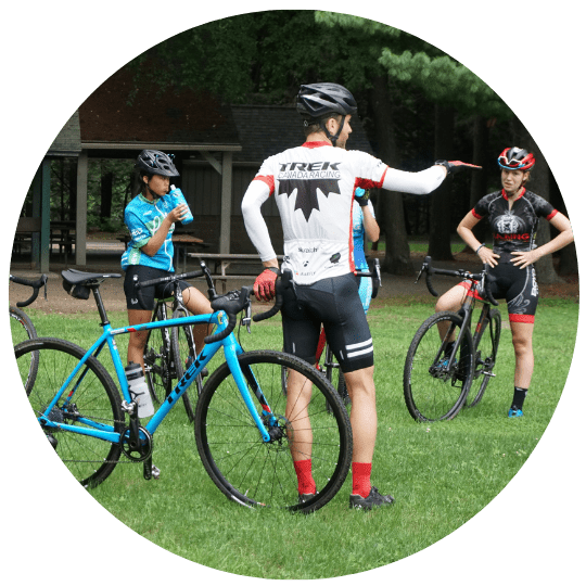A group of cyclists in cycling gear stands with their bicycles on a grassy area. One cyclist with a white and red jersey is gesturing with his hand while others listen. Another cyclist wearing a blue jersey is drinking from a bottle. Trees and structures are in the background.