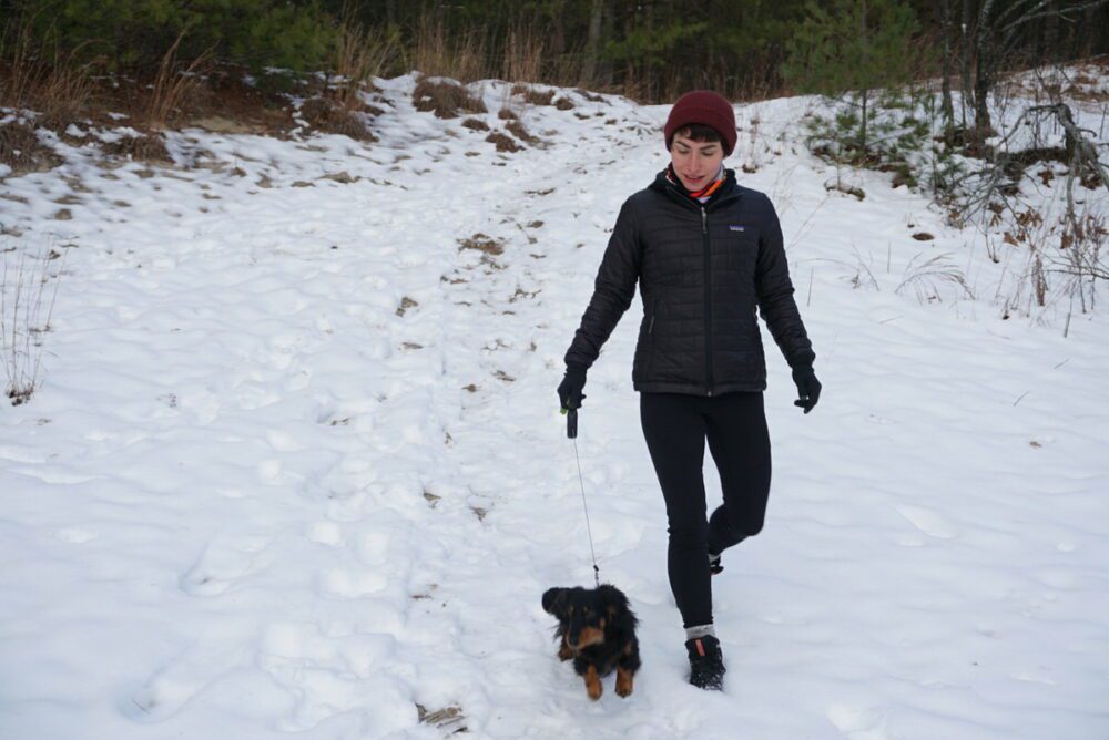 A person wearing a black jacket, black pants, and a red beanie is walking a small black and brown dog on a leash in a snowy outdoor setting. Trees and sparse vegetation are in the background.