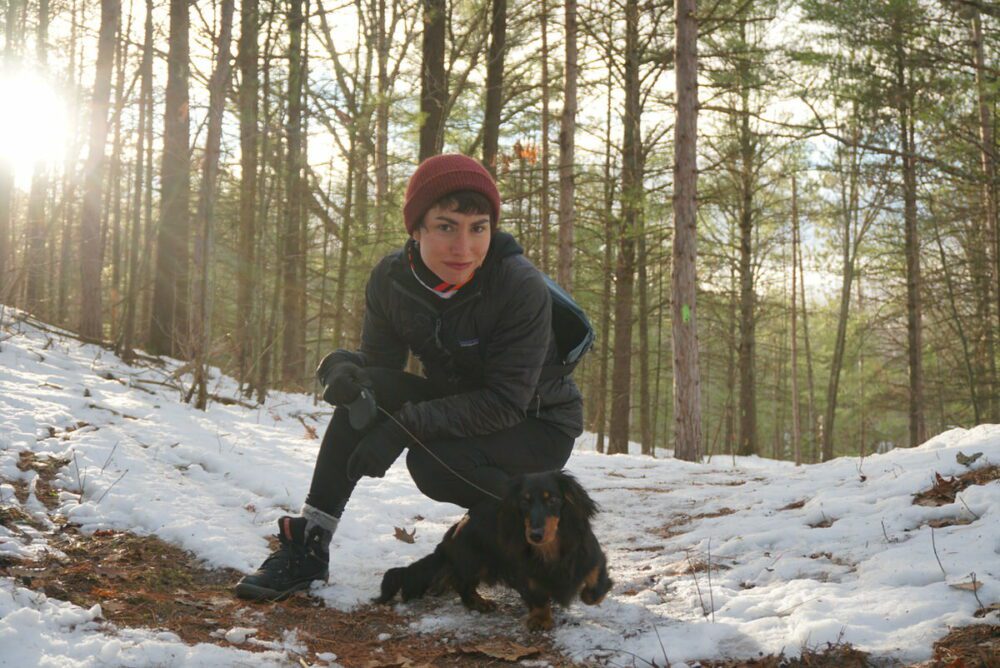 A person dressed in winter clothing crouches on a snowy forest path, holding a leash attached to a small black dog. The sun is shining through the trees in the background.