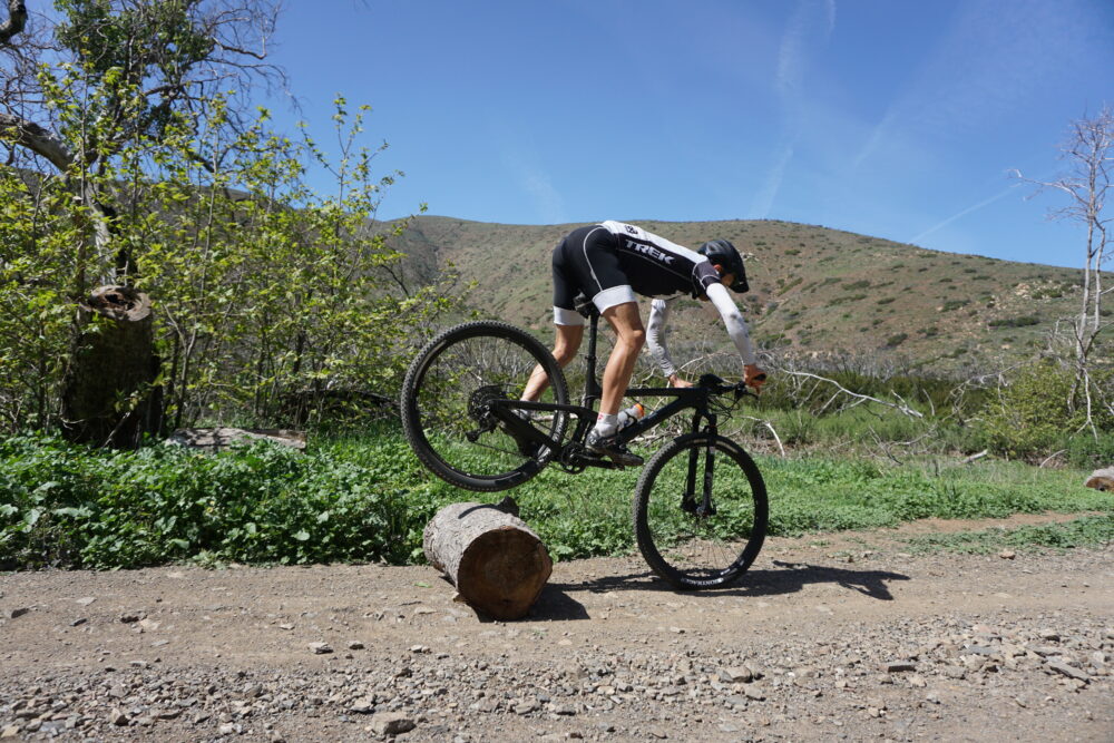 A cyclist navigates a mountain bike over a log obstacle on a dirt trail surrounded by green foliage and hills under a clear blue sky. The rider is balancing the front wheel on the log while lifting the rear wheel off the ground.