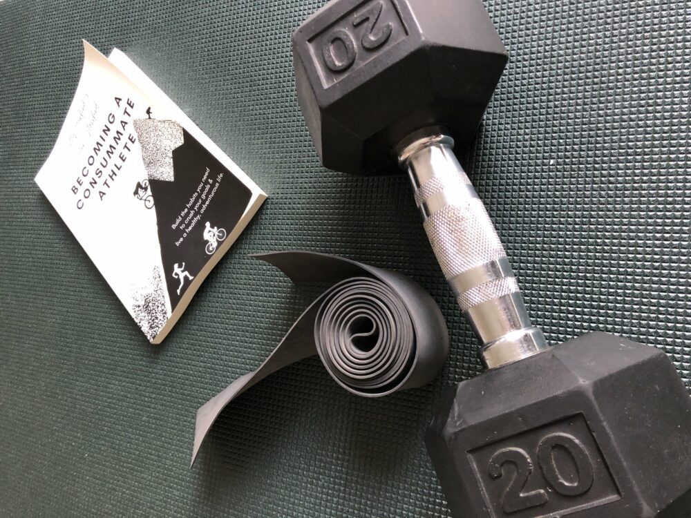 A 20-pound black dumbbell lies next to an unrolled yoga mat and an open book titled "becoming a supple leopard" on a green textured surface.