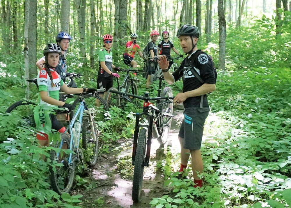 A group of seven cyclists, wearing helmets and cycling gear, are stopped on a forest trail with their mountain bikes. One cyclist in the foreground, wearing a black shirt, is gesturing, while the others stand or sit on their bikes among dense greenery.