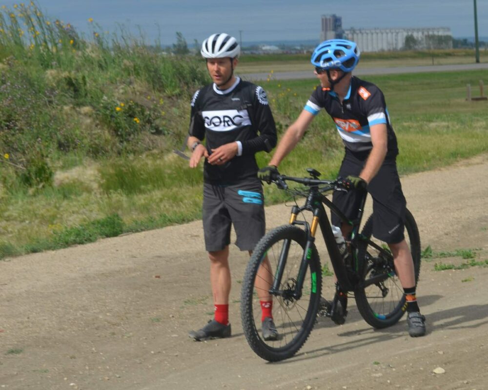 Two cyclists in cycling gear, including helmets and gloves, stand next to their bicycles on a dirt path. They appear to be having a conversation. The backdrop includes grassy areas, bushes, and a distant industrial building.