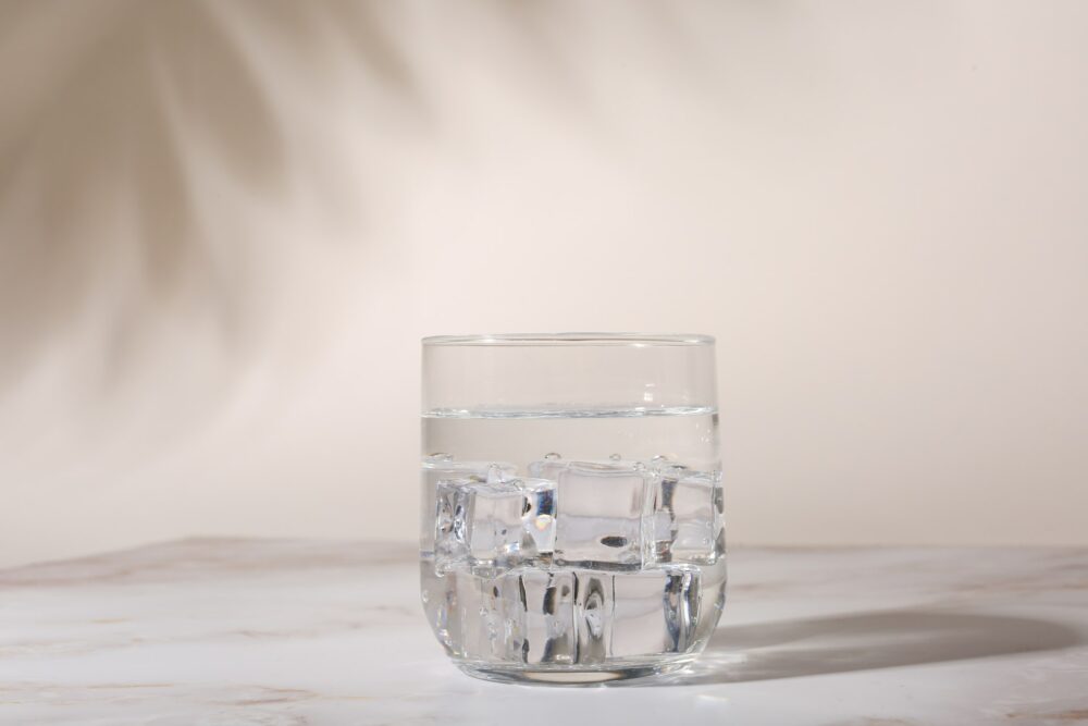 A clear glass filled with water and ice cubes sits on a light-colored surface. Shadows and soft lighting are cast in the background, creating a serene and minimalist atmosphere.