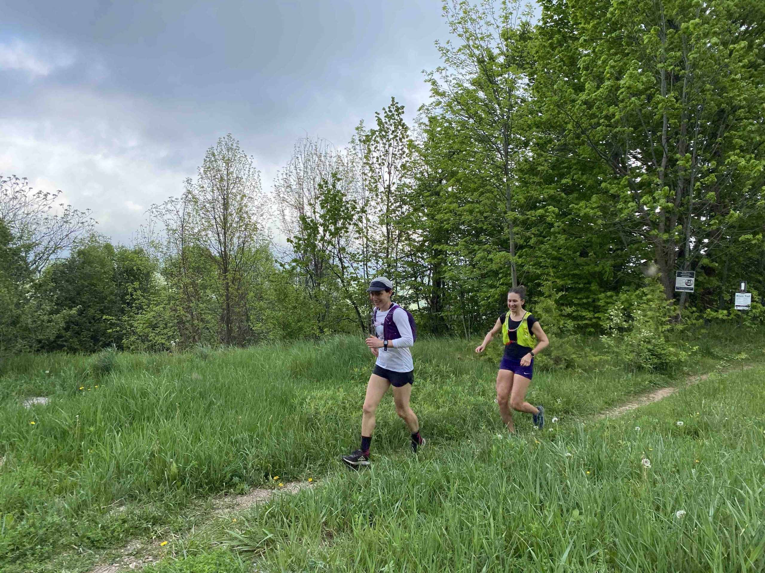 Two people are running on a grassy trail through a forested area. The runner on the left wears a white shirt, black shorts, and a hat. The runner on the right wears a yellow tank top and blue shorts. Trees and a cloudy sky are in the background.