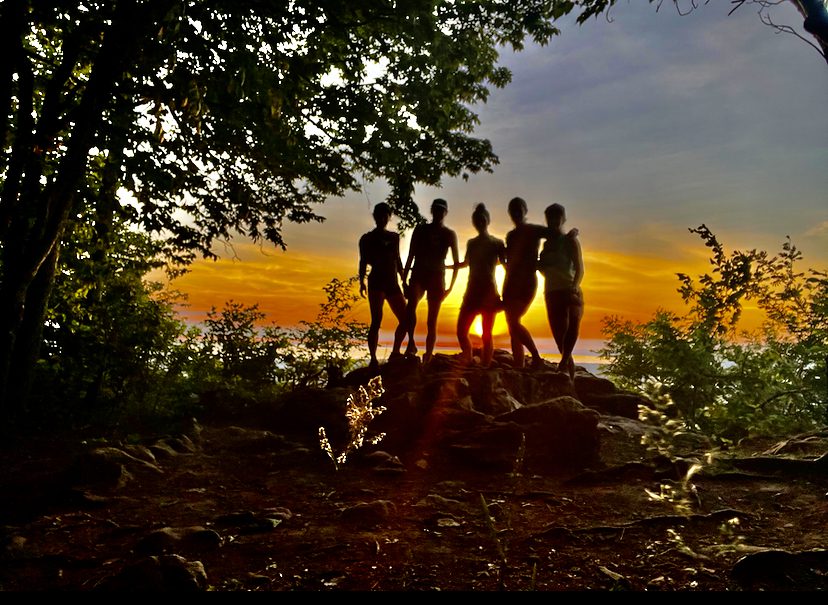 Five people stand on a rocky outcrop surrounded by trees, silhouetted against a vibrant sunset. The sky transitions from orange to more muted tones near the horizon. The foreground is shaded, and the ground appears uneven with scattered rocks and foliage.