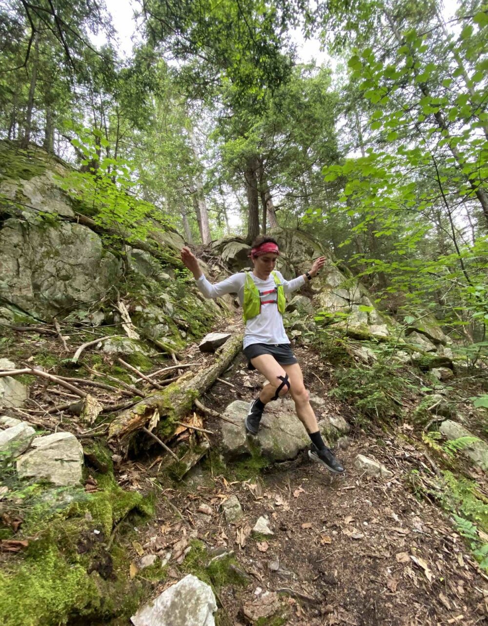 A person wearing a white shirt, green vest, and black shorts is mid-jump on a rocky forest trail. They have a red headband and black knee brace, and the background consists of trees, shrubs, and rocks. The person appears to be trail running or hiking.