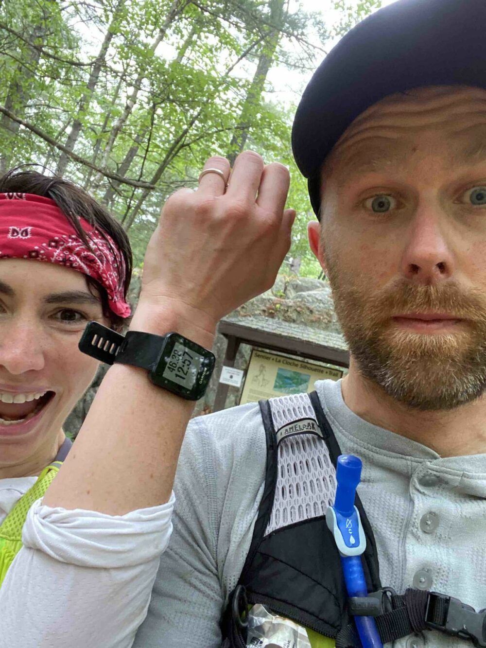 Two individuals, one wearing a pink bandana and the other a white cap, pose outdoors in a wooded area. The person on the right sports a hydration pack and raises a hand to show a smartwatch. Both appear to be dressed in athletic gear for an outdoor activity.