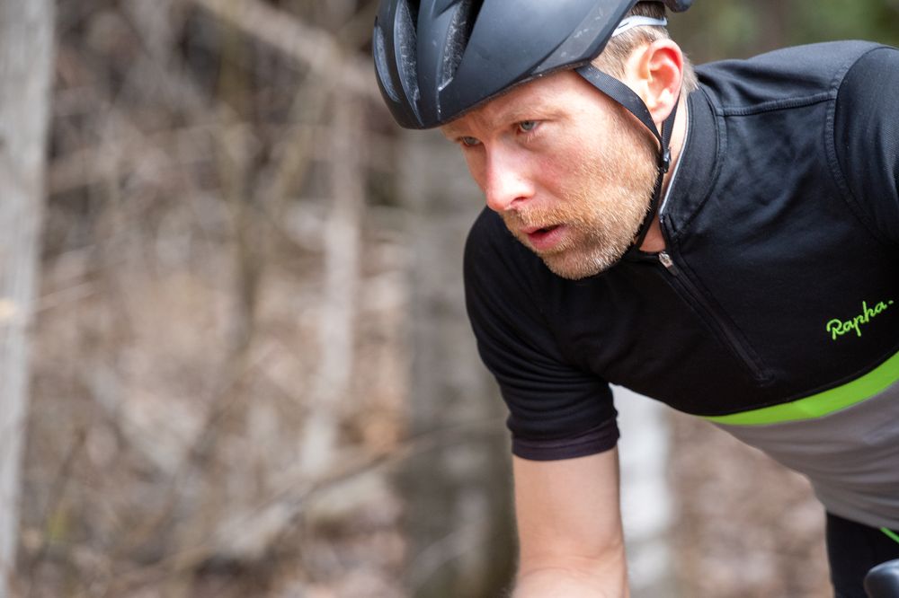 A cyclist wearing a black helmet and a black cycling jersey with green accents is focused and pedaling. The background appears to be an outdoor, wooded area.