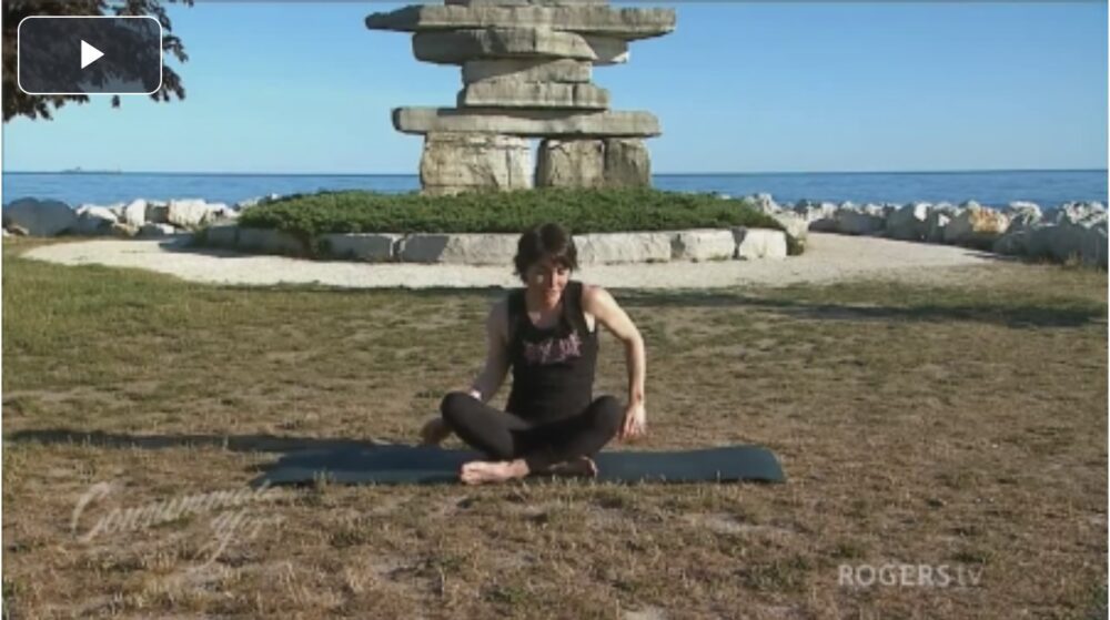 A person sits cross-legged on a yoga mat in a grassy area near a lake. Behind them is a large stone sculpture and a clear, blue sky. The image appears to be paused from a video, with the text "Community" and "ROGERS TV" visible on the bottom.