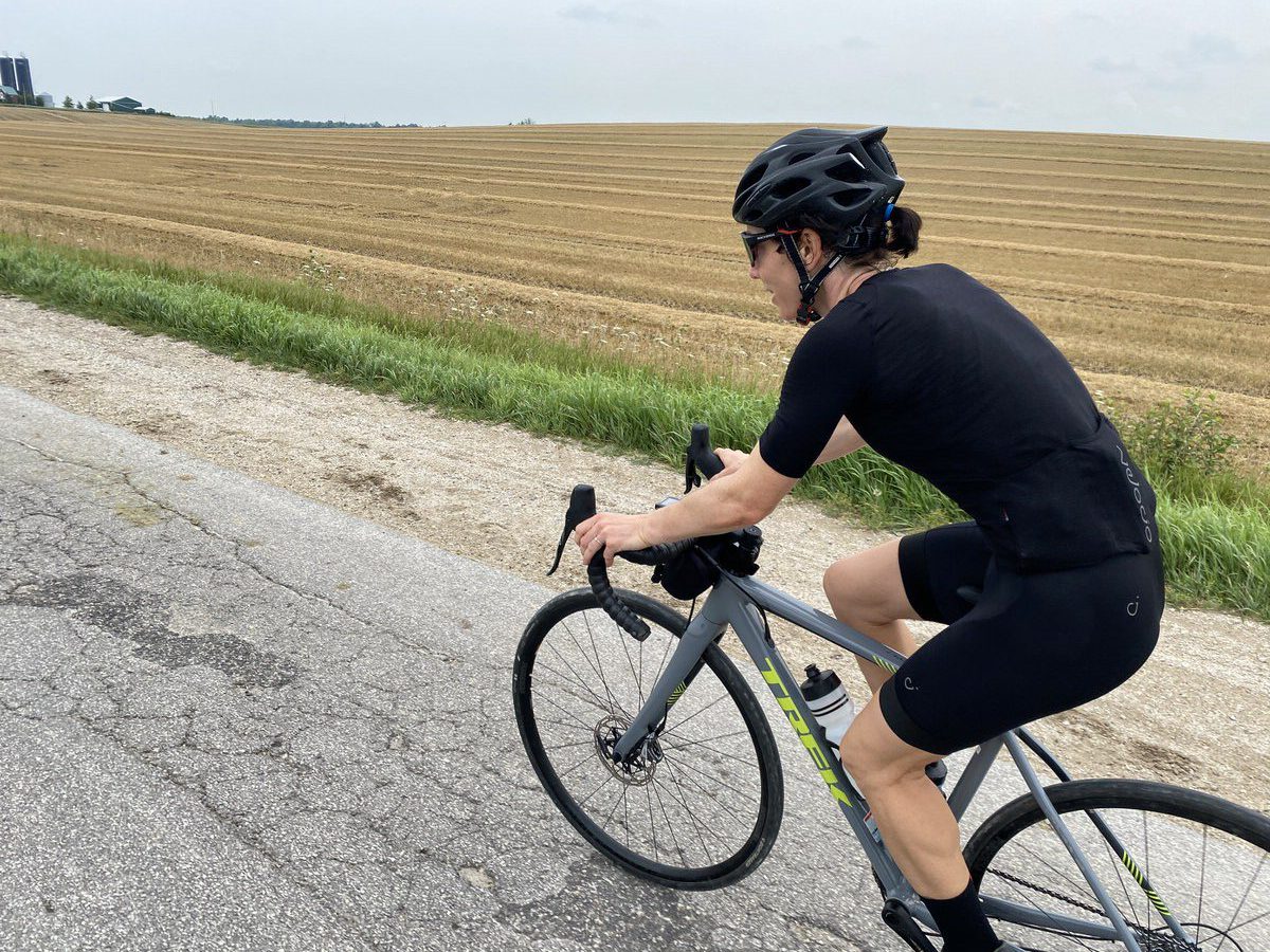 A cyclist in black attire and a helmet rides a road bike on a paved surface adjacent to a plowed field. The cyclist is positioned in mid-pedal, leaning forward, and grasping the handlebars. The sky above is clear with minimal clouds.