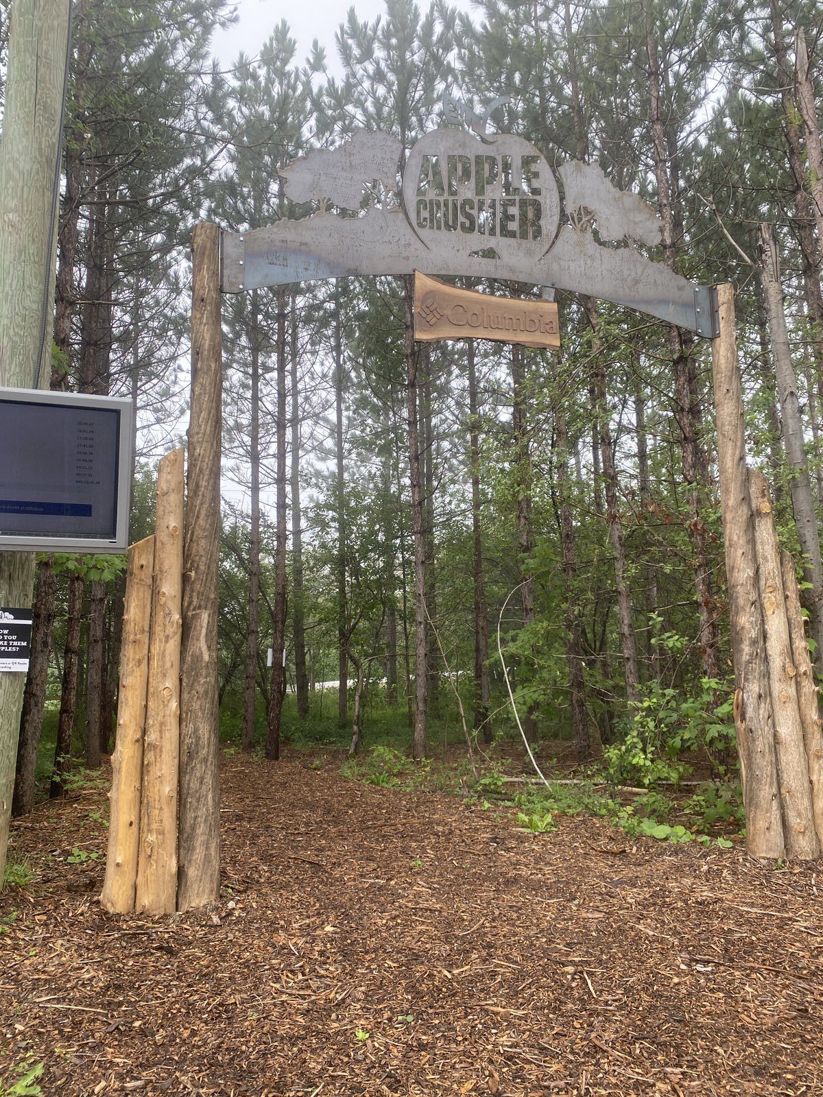 A rustic wooden archway labeled "Maple Crusher" marks the entrance to a forest trail. Behind the arch, tall trees line the path, and a small sign is visible to the right. The ground is covered in wood chips, and an information board stands to the left of the entrance.