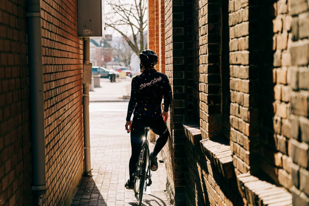 A person wearing cycling gear and a helmet rides a bike through a narrow alley between two brick buildings. Sunlight illuminates the alley and casts shadows, while the background shows an open street.