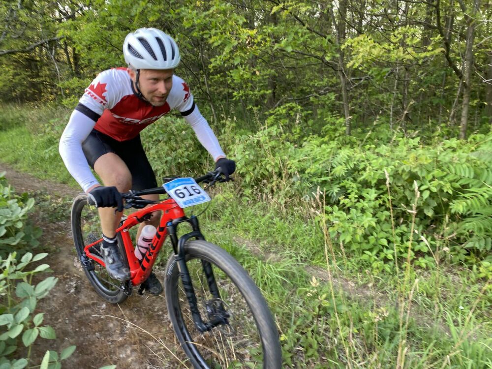 A cyclist in a red and white jersey with the number 616 attached rides a mountain bike along a dirt trail surrounded by greenery. The rider is wearing a white helmet and black shorts and is focused on the path ahead.