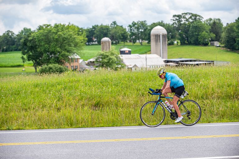 A cyclist wearing a blue jersey and yellow helmet rides a road bike on a rural road. In the background, there is a grassy field, farm buildings, including silos, and trees under a partly cloudy sky.
