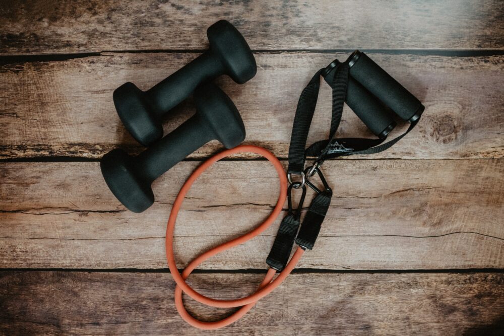A pair of black dumbbells and an orange resistance band with black handles are placed on a wooden surface. The equipment is arranged side by side, suggesting a fitness or workout setting.