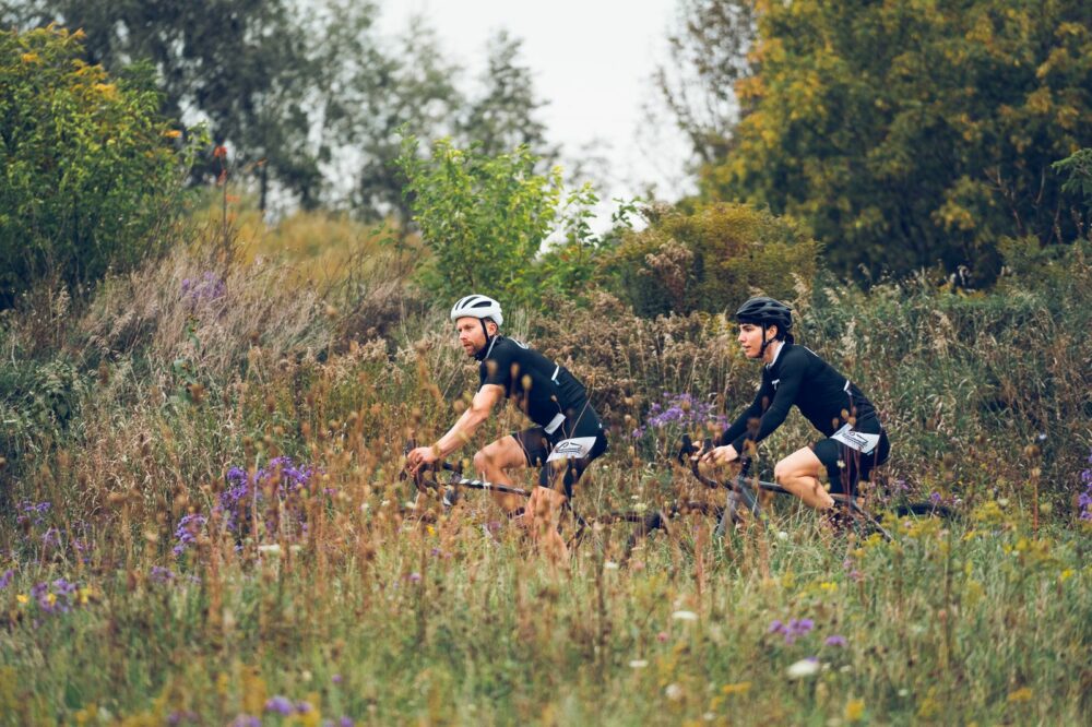 Two cyclists are riding mountain bikes through a grassy and slightly overgrown trail surrounded by tall plants and trees. Both cyclists are wearing black cycling outfits and helmets, and the background features a mix of greenery and wildflowers.