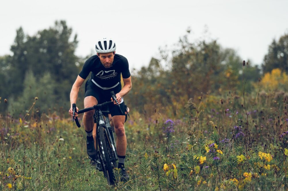 A cyclist wearing a black outfit and white helmet rides a bicycle through a grassy and flower-filled terrain. Trees and foliage are visible in the background. The cyclist appears to be focused and determined.