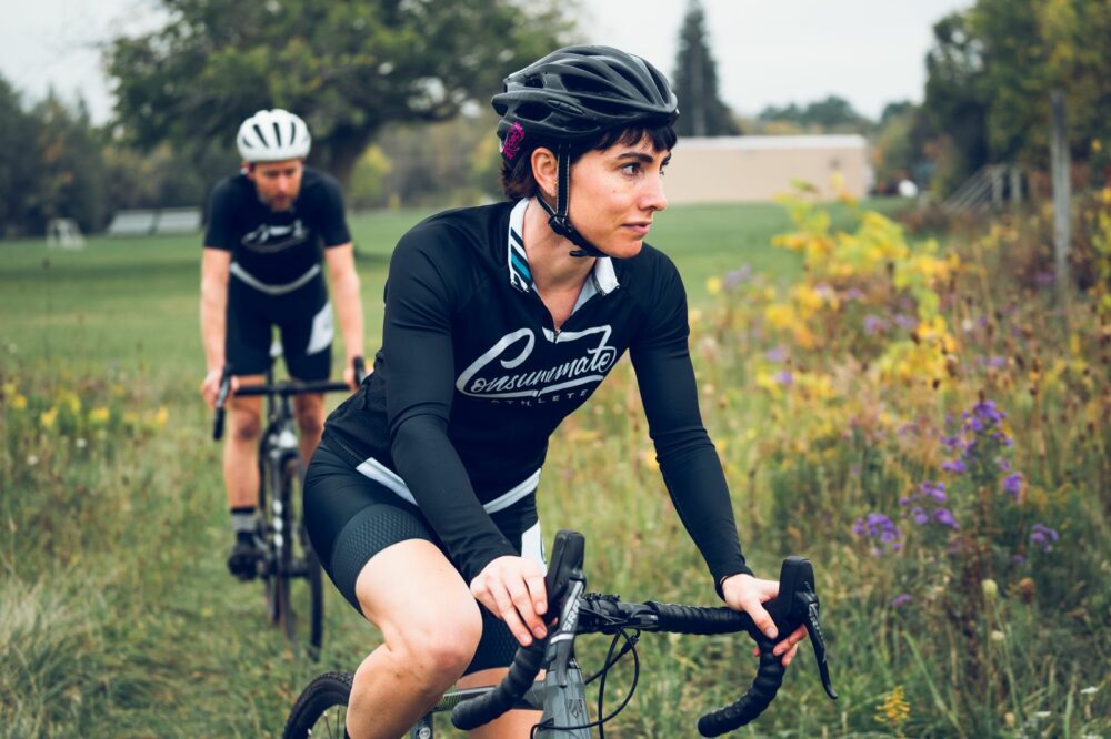 Two cyclists wearing helmets and black cycling jerseys are riding on a path through a grassy and wooded area. The cyclist in the foreground is focused ahead while the one in the background follows at a short distance. The day appears overcast.