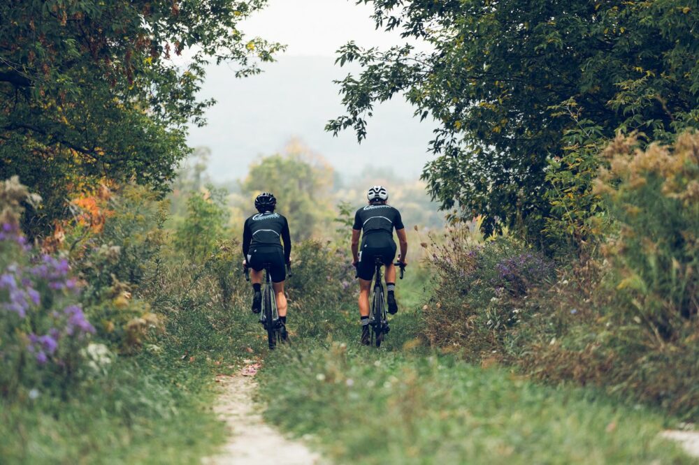 Two cyclists in black gear ride on a narrow, dirt trail through a densely wooded area. Surrounding foliage is lush and green, with some purple flowers visible. The sky is overcast, and the trail appears to stretch into the distance ahead of them.