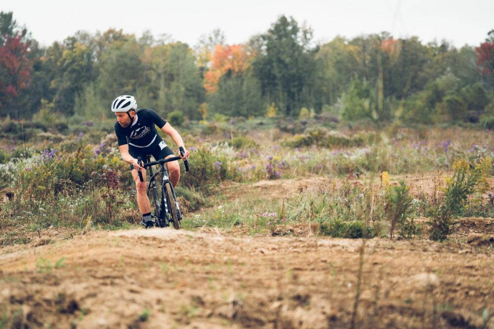 A person wearing a black cycling outfit and helmet rides a bike on a dirt trail through an open field with scattered patches of wildflowers. Trees with autumn foliage are visible in the background under a cloudy sky.