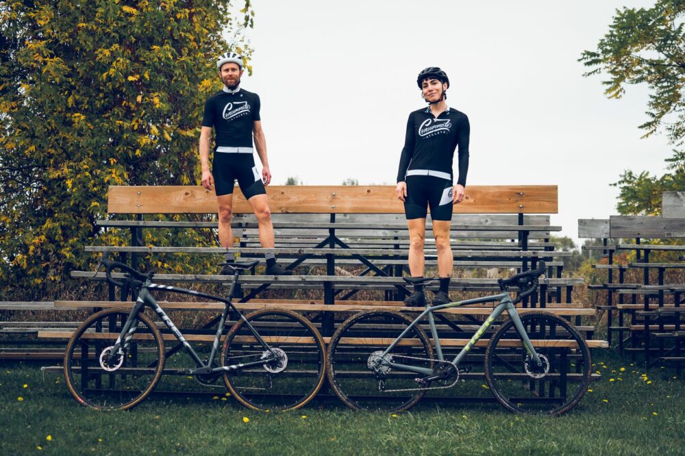 Two cyclists in black cycling outfits stand on a wooden bleacher near two bicycles. Both are wearing helmets, and one is slightly taller than the other. There are trees with green and yellowing leaves in the background, and the grass has some scattered dandelions.