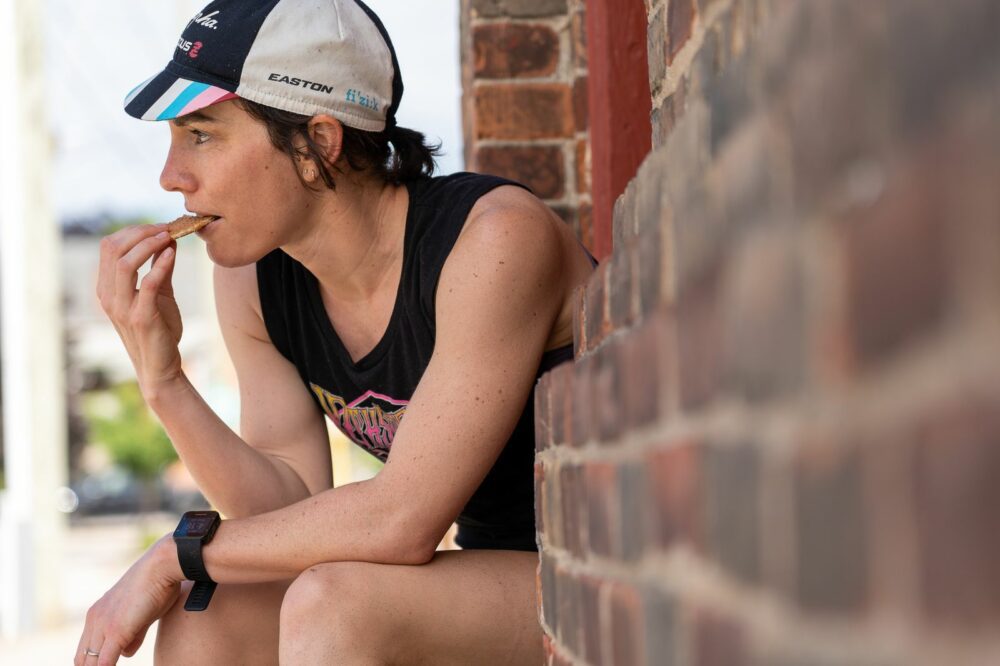 A person wearing a cap and a wristwatch is sitting outside next to a brick wall, eating a snack. They are wearing a sleeveless black shirt and looking towards the left, appearing thoughtful.