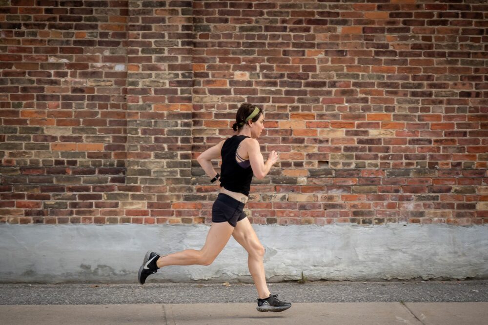 A person wearing a black tank top, black shorts, and black shoes is running along a sidewalk in front of a brick wall. They have a green headband and are in profile view, mid-stride, with their left foot off the ground.