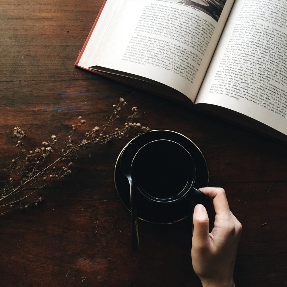 A hand holding a small black cup of coffee next to an open book on a wooden table. Dry twigs with small flowers are placed diagonally across the table beside the cup and book. The scene suggests a quiet, contemplative moment.