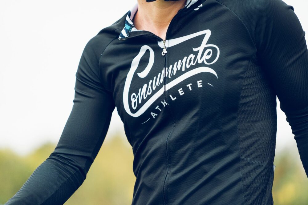 Close-up of a person wearing a black athletic jacket with the words "Consummate Athlete" printed on the front in white script. The image shows the upper part of the person's body, focusing on the jacket's text. The background is blurred natural scenery.