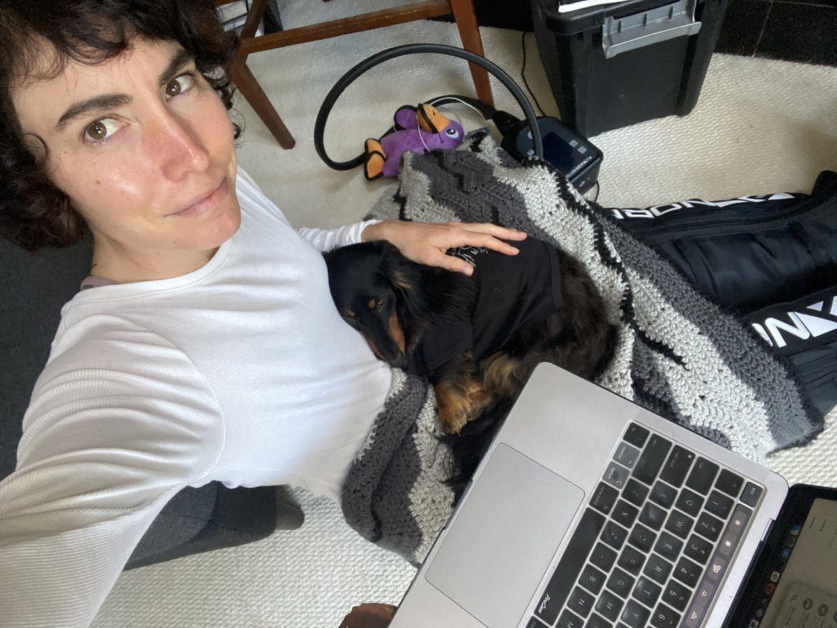 A woman sits on the floor with a laptop on her lap and holds a small black dog wrapped in a gray blanket. There's an exercise bike and a barbell nearby, suggesting a home office and workout space.