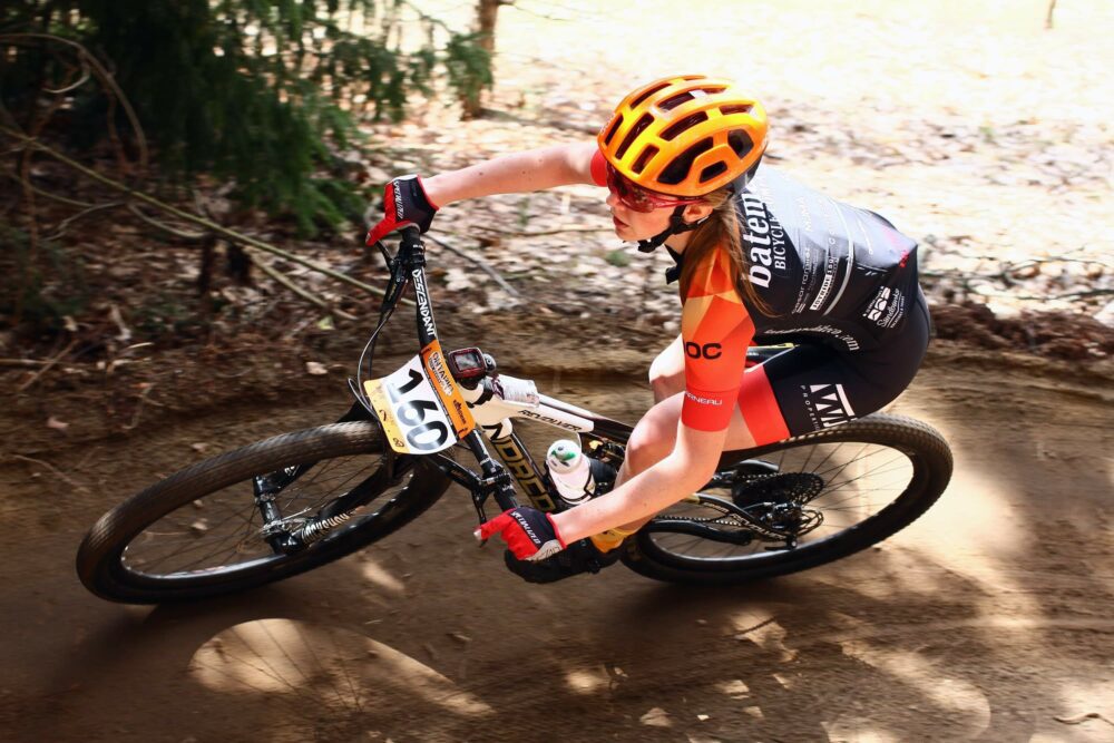 A cyclist wearing an orange helmet and black cycling jersey rides a mountain bike around a dirt trail curve. The cyclist's number plate reads "306." Trees and foliage are visible in the background.