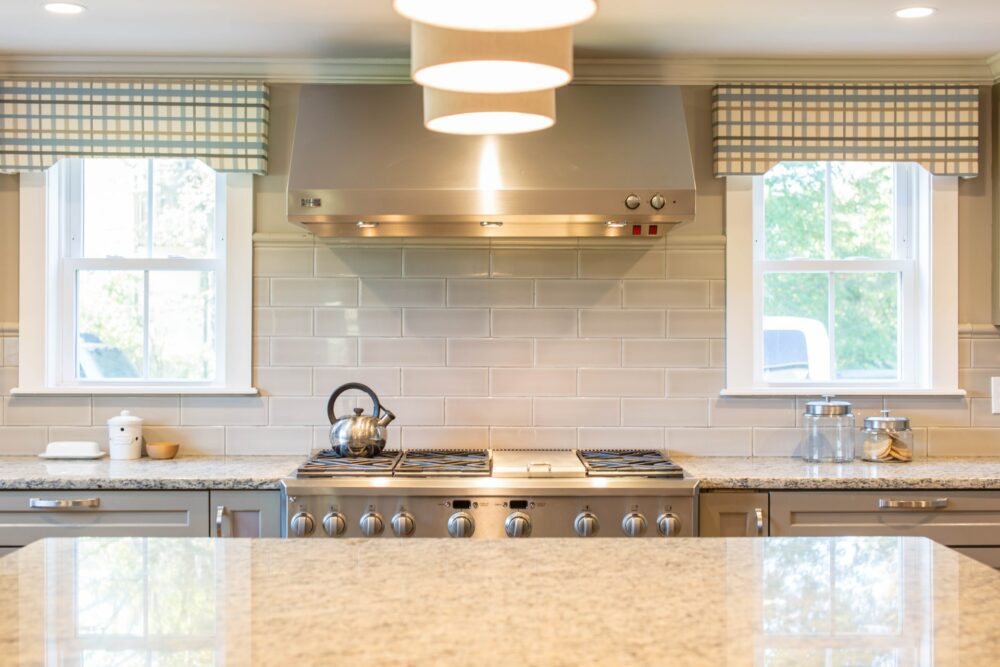 A modern kitchen with a granite countertop and stainless steel appliances. Two large windows with checkered valances are above the stove, which has a kettle on one of the burners. The backsplash features light-colored subway tiles, and a hanging light fixture is visible.