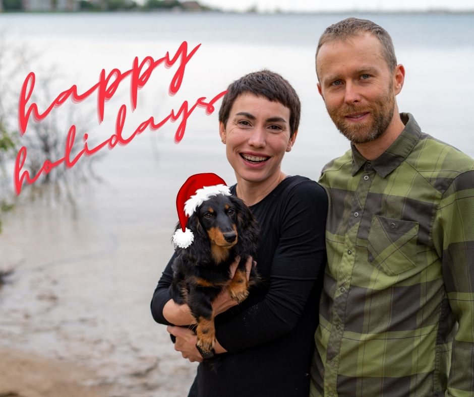 A woman and man stand by a lakeside, smiling at the camera. The woman is holding a small black and brown dog wearing a Santa hat. The words "Happy Holidays" are written in red script in the upper left corner of the image.
