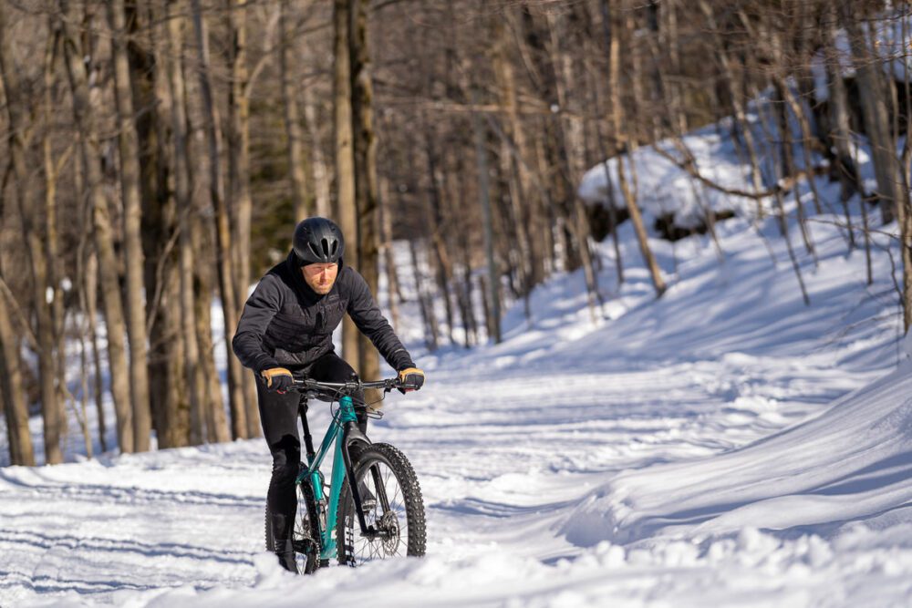 A person wearing a black helmet and winter clothing is riding a teal-colored fat bike on a snow-covered trail in a forested area. The surroundings are filled with leafless trees and snow.