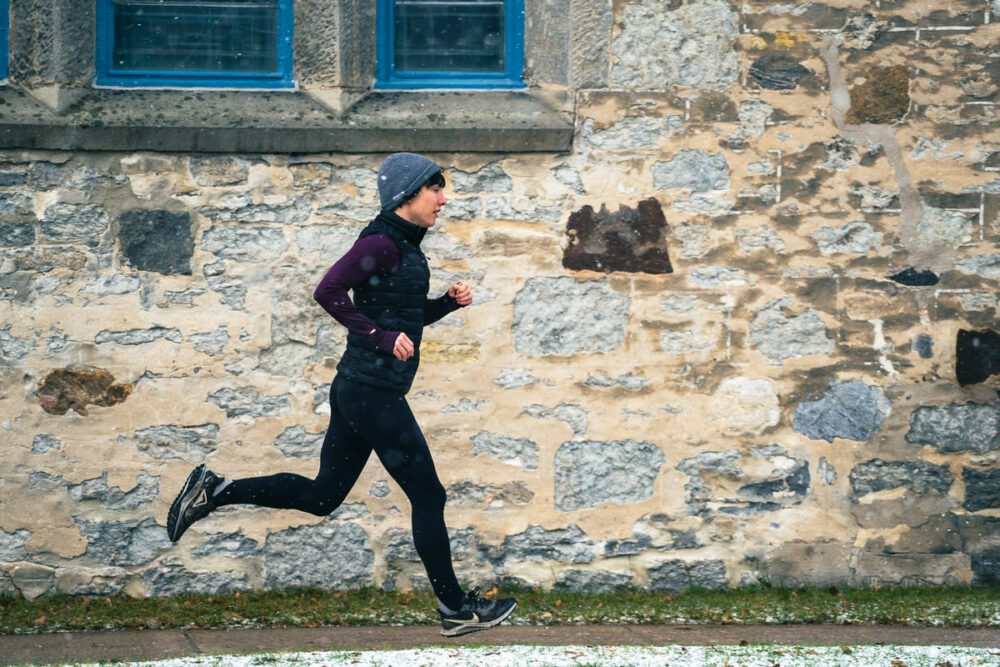 A person in athletic clothing and a beanie is running beside a stone wall. The ground shows patches of snow, suggesting a cold weather setting. The wall includes two windows with blue frames.